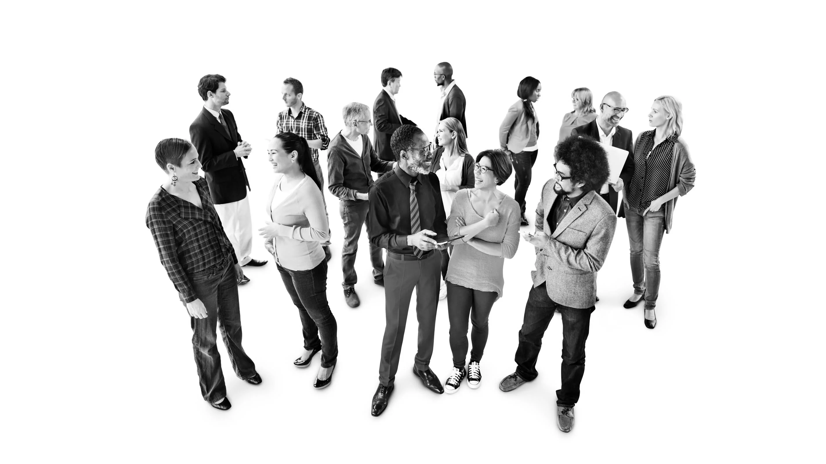 A diverse group of approximately fifteen people of various ages and styles stand together against a white background in a black and white photograph, some conversing in small clusters, others standing independently.