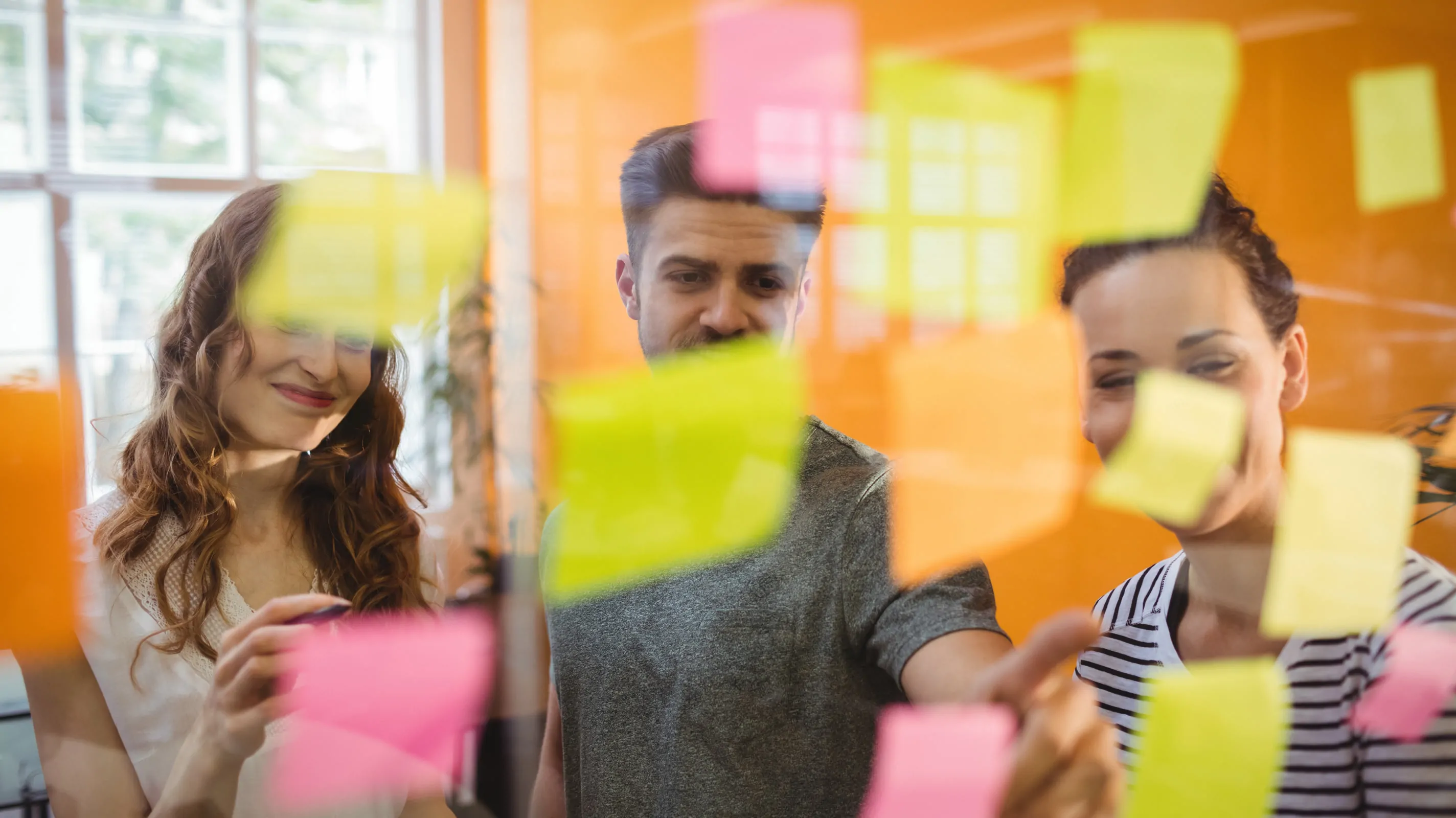 Three team members organize colorful sticky notes on a glass wall during a collaborative affinity mapping session.