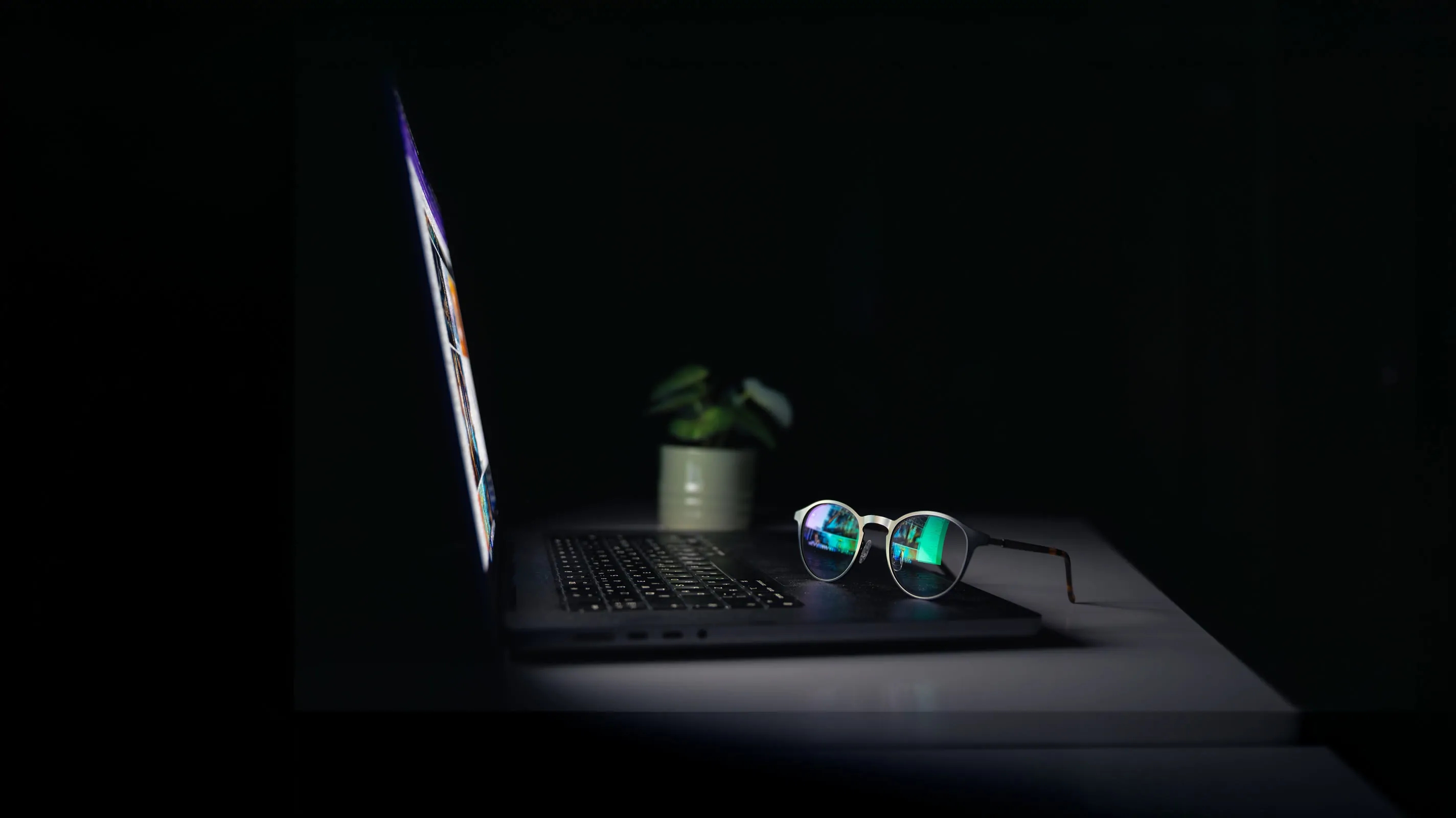 A pair of glasses rests on a laptop keyboard in a darkened room, with the screen casting a faint glow and a small potted plant visible in the background.
