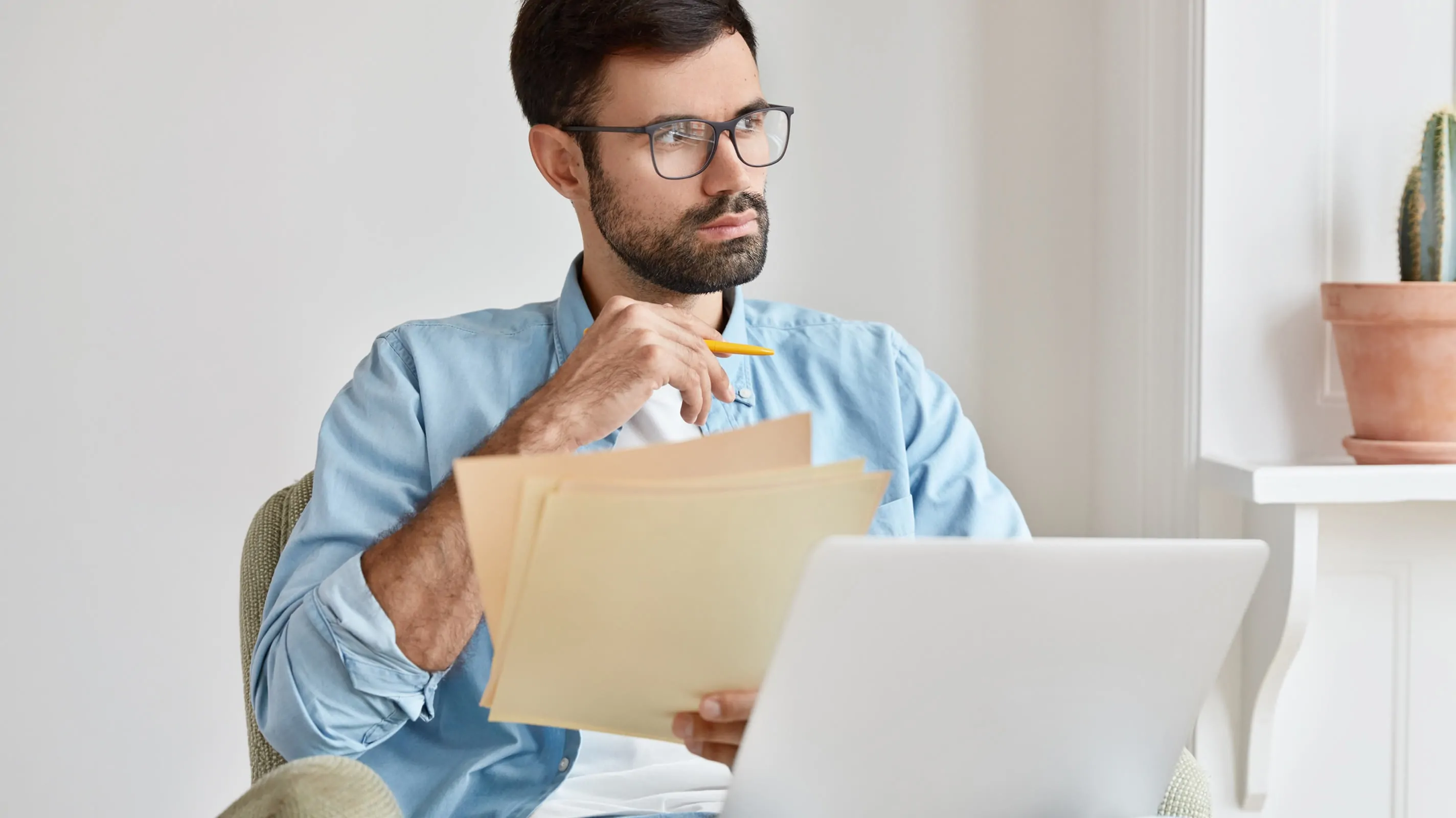 A man wearing glasses and a blue shirt sits at a desk holding a folder of documents and a pencil, gazing thoughtfully to the side. An open laptop is in front of him and a small cactus sits on the windowsill behind him.