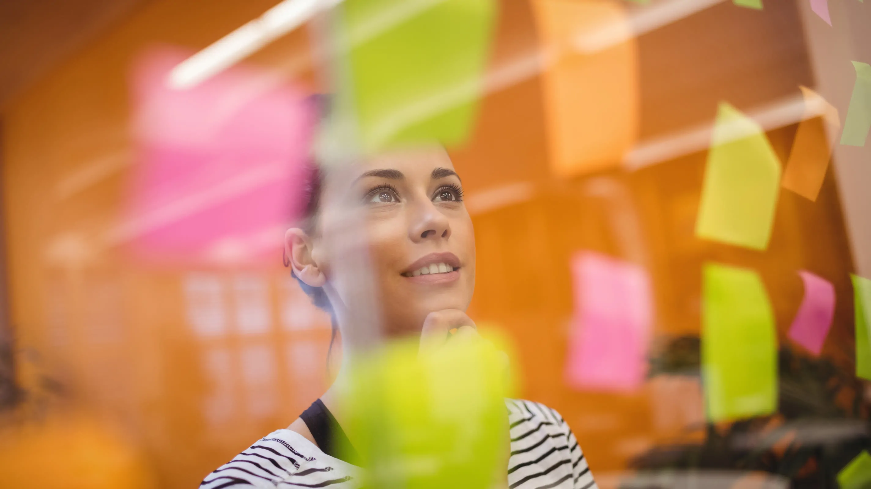 A woman stands behind a glass wall covered in colorful sticky notes — pink, green, and orange — in what appears to be a design or research workshop setting. She looks upward thoughtfully, resting her chin on her hand.