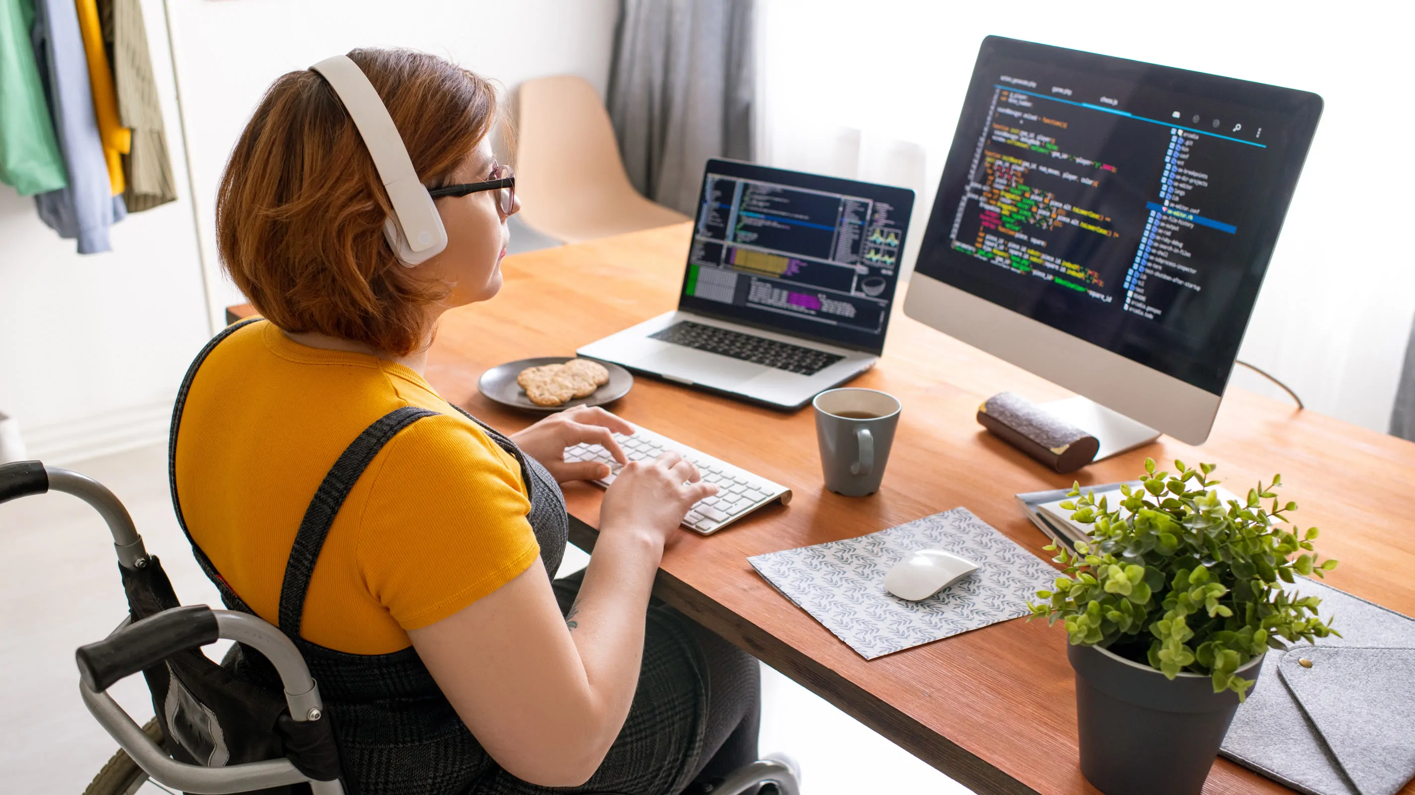 A young woman using a wheelchair sits at a wooden desk, wearing headphones and glasses, typing on a keyboard while working across a laptop and an external monitor both displaying syntax-highlighted code.