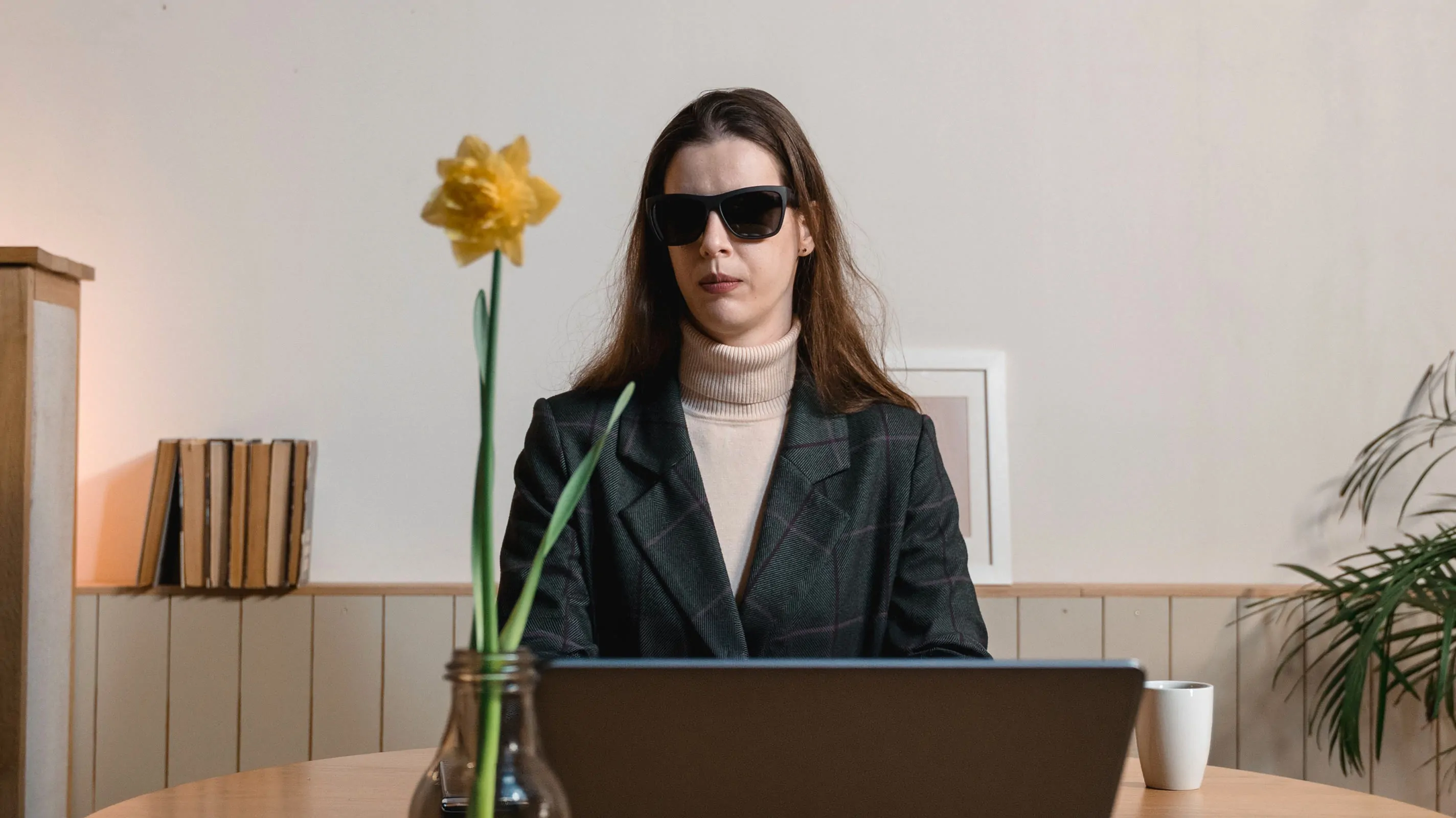 A woman wearing dark opaque sunglasses sits at a wooden desk in front of a laptop, suggesting visual impairment. A yellow daffodil in a glass vase, a white cup, and a row of books are visible in the warm, minimal workspace around her.