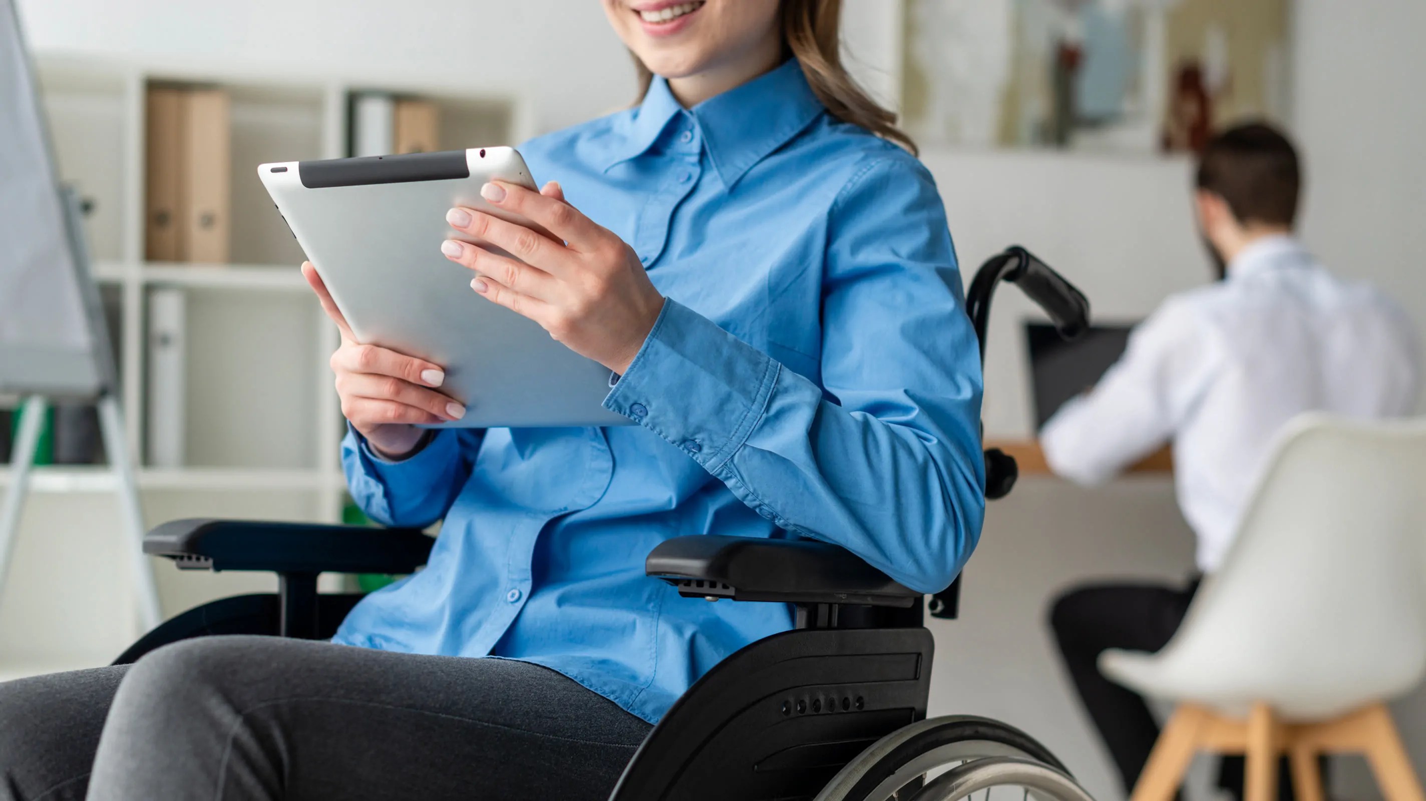 A woman who uses a wheelchair works on a tablet in an office, with a colleague working at a desk in the background.