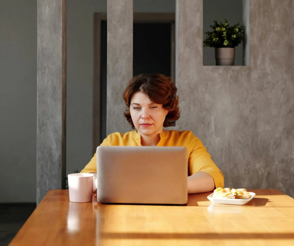 A woman focuses on her laptop at a kitchen table, with a coffee mug and snack beside her.