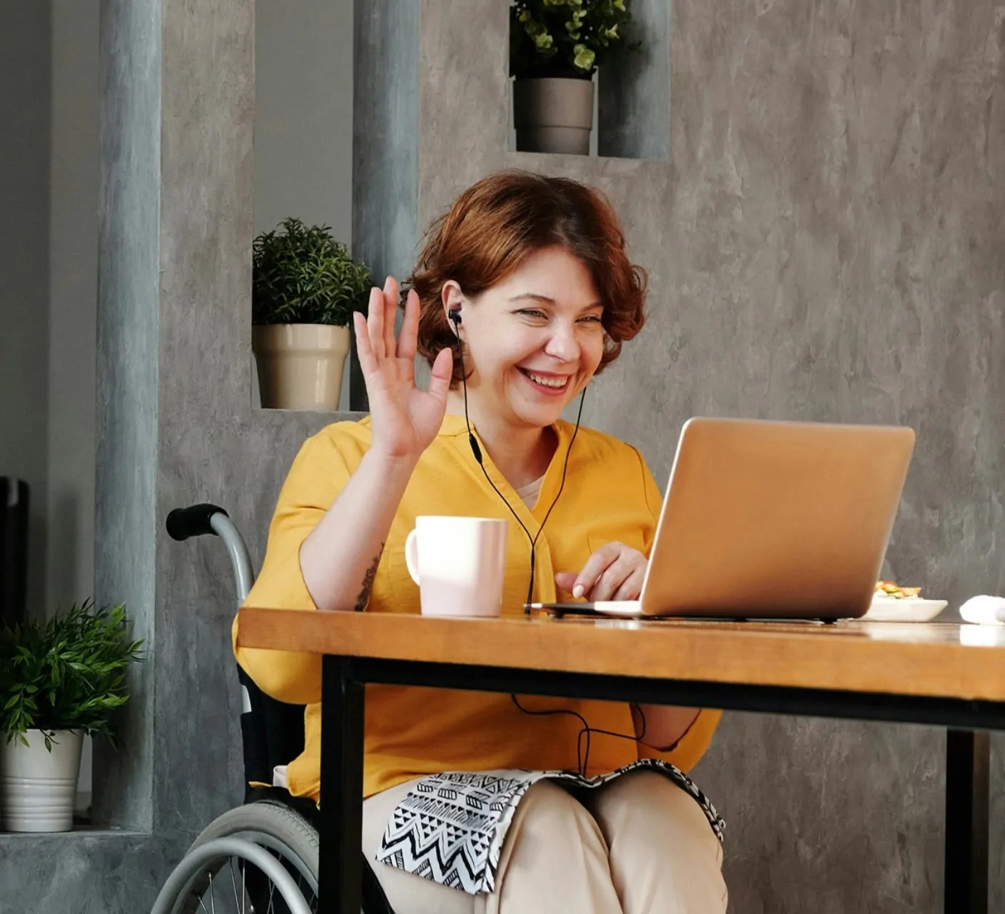 A woman who uses a wheelchair waves at her laptop screen during a video call, smiling and wearing earphones.