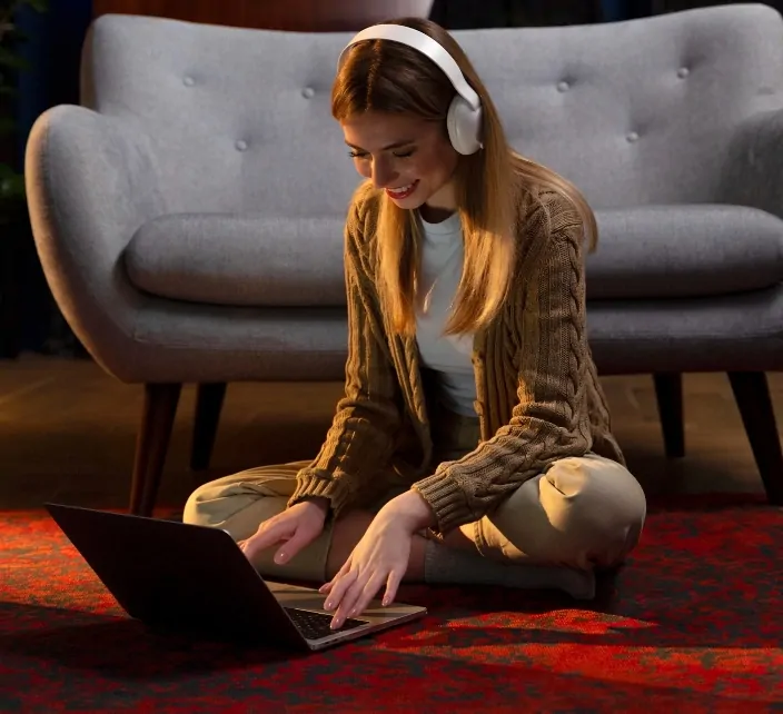 A woman wearing headphones sits cross-legged on the floor, using a laptop to participate in an online session.