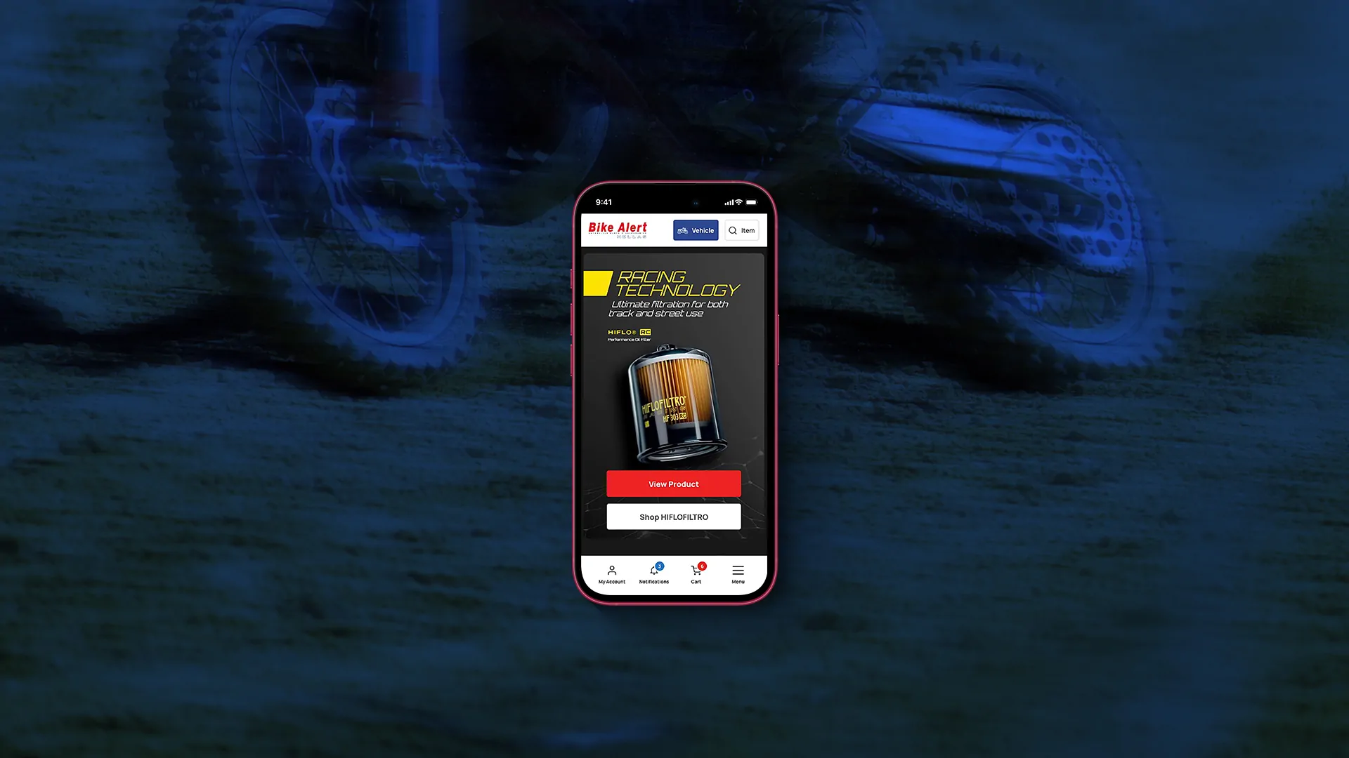 A smartphone displaying the Bike Alert Hellas mobile homepage with its "Racing Technology" hero banner for a performance oil filter, set against a stylized blue-toned background image of a motocross rider's wheel and chain.