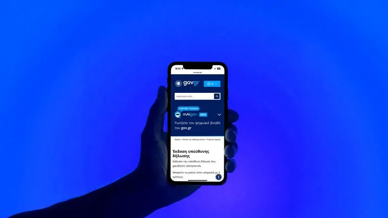 A hand holds a smartphone displaying the gov.gr mobile website, showing the mAIgov AI assistant and a solemn declaration service page, against a blue background.