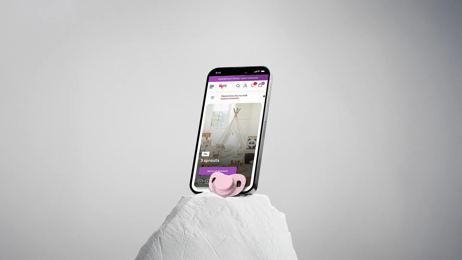 A smartphone displaying the irokids.gr mobile e-commerce homepage, showing a children's product collection banner, propped on a stone surface next to a pink pacifier.