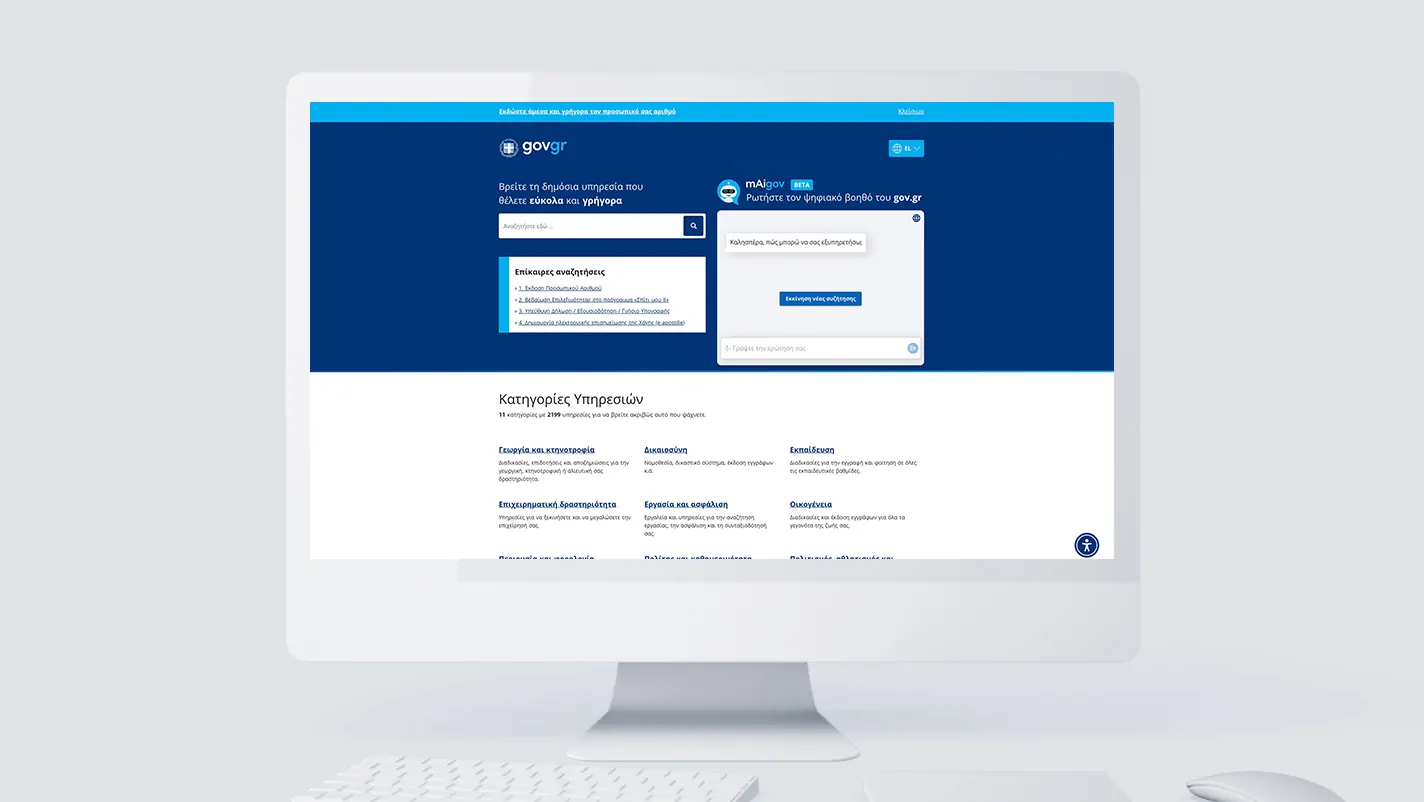 A desktop monitor displaying the gov.gr homepage, showing the main search interface, the mAIgov AI assistant panel, and a service categories grid below.