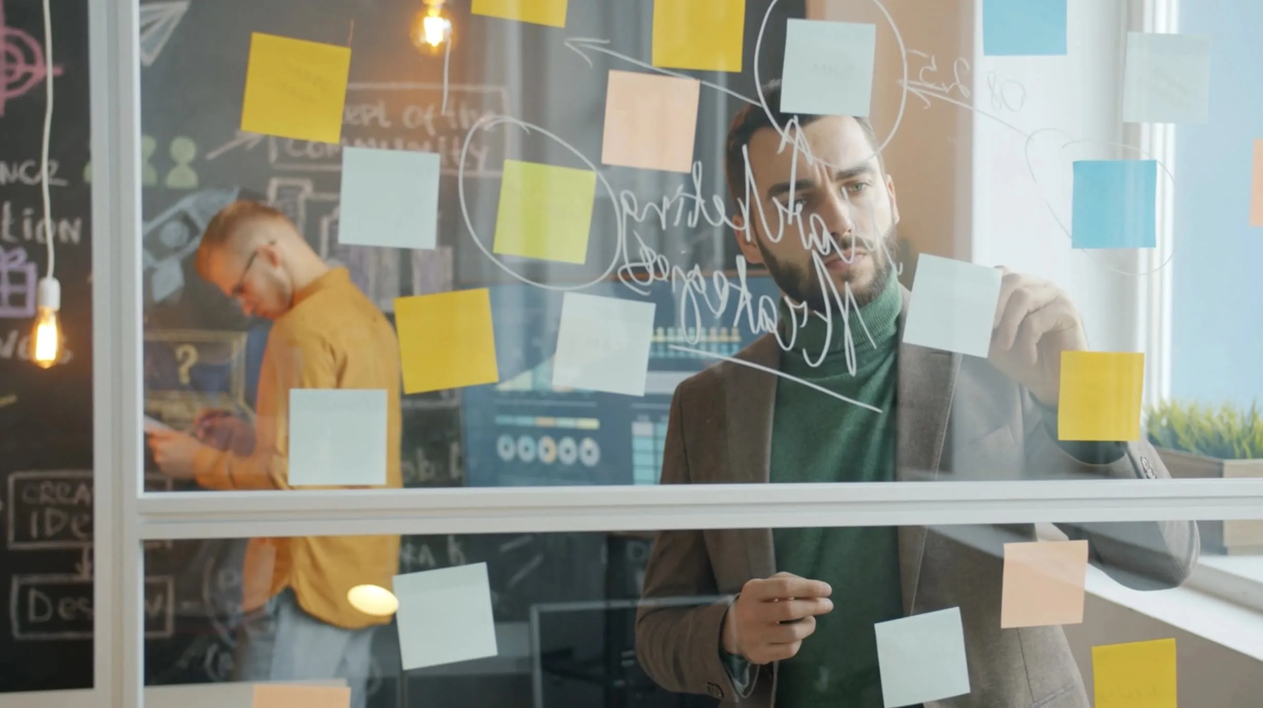 Two people collaborate in a workshop, writing on a glass wall covered with sticky notes and handwritten diagrams.