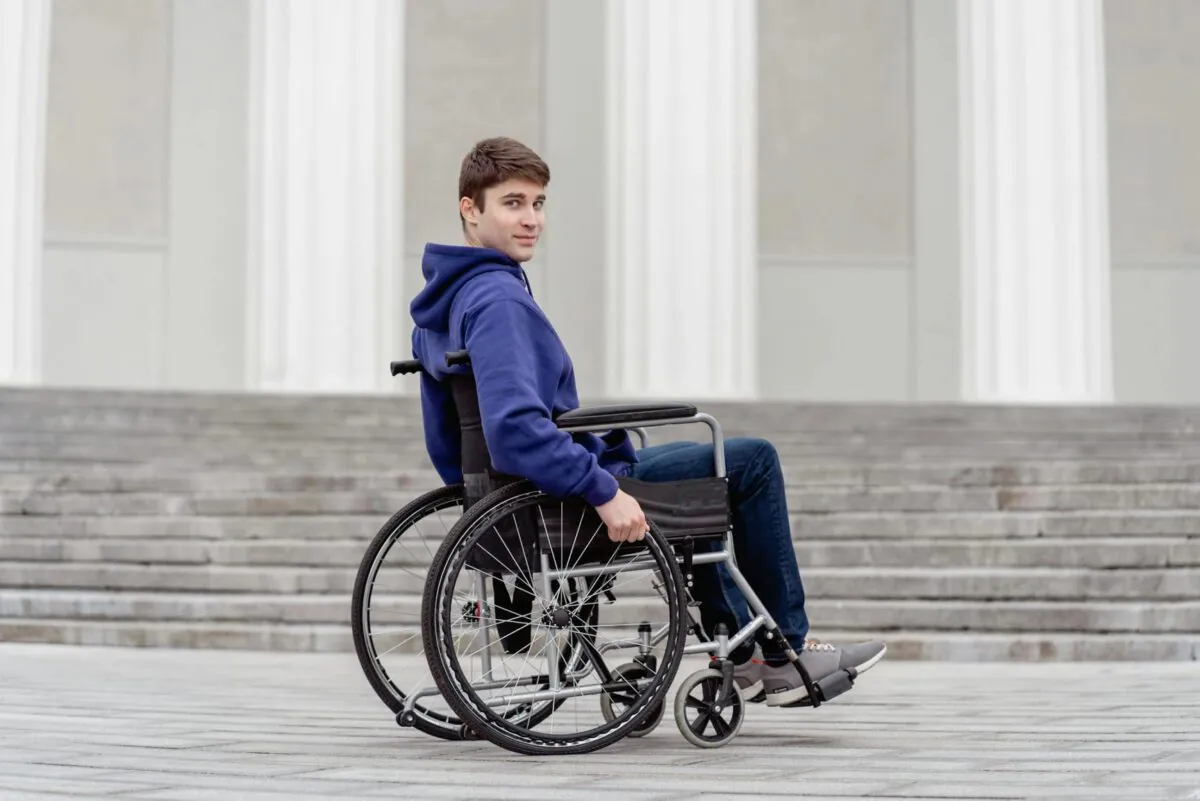 A young man in a wheelchair looks over his shoulder, positioned in front of a building with wide steps.