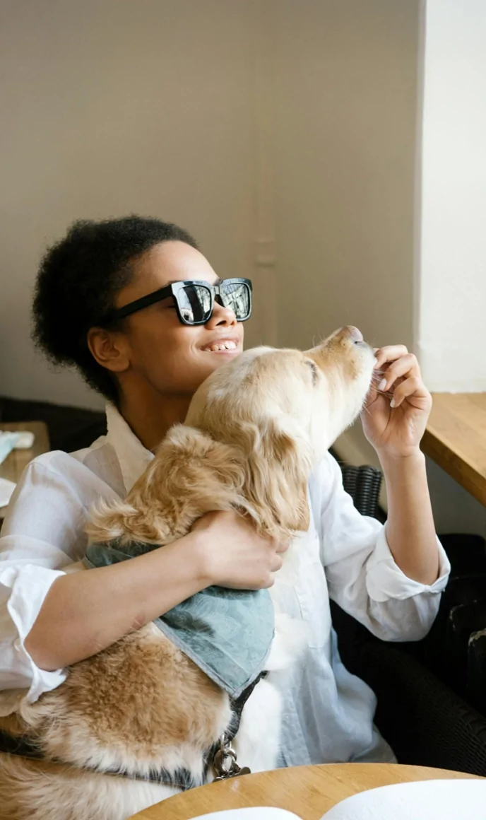 A blind woman smiles while her guide dog nuzzles close to her.
