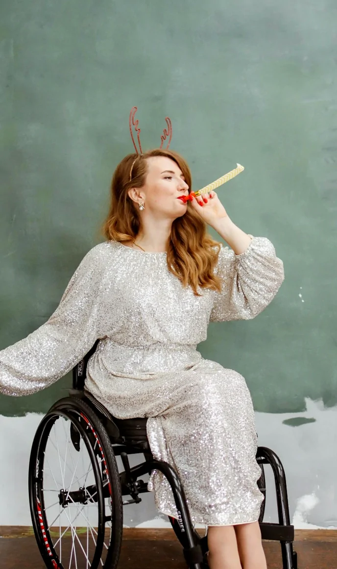 A woman in a wheelchair wearing a festive outfit blows a party horn, smiling confidently.