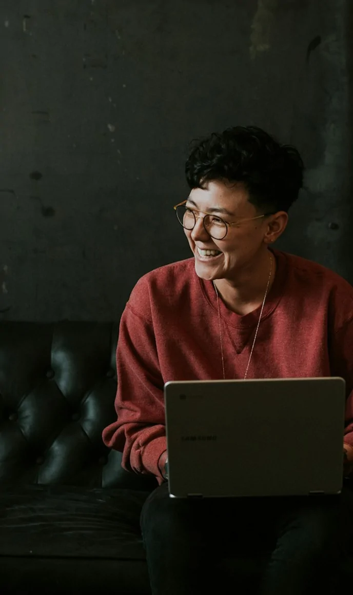 A woman laughs while working on a laptop, seated on a sofa.