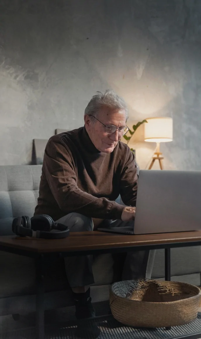 An older man works focused on a laptop in a dimly lit living room.