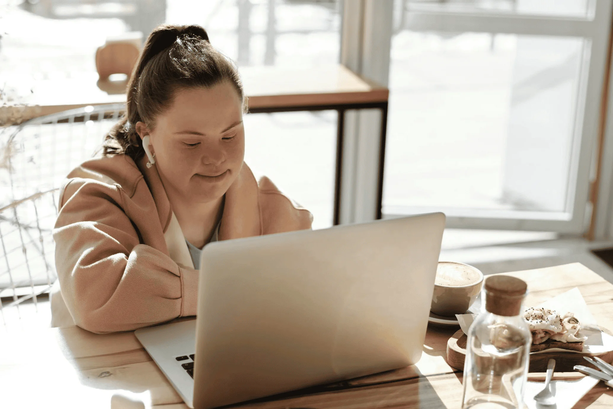 A young woman with Down syndrome smiles while using a laptop at a café table.