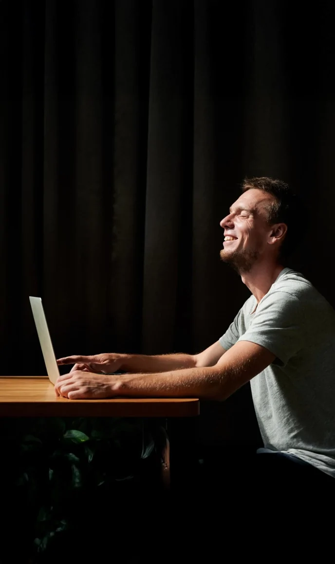 A man sits at a desk with a laptop, smiling and looking upward, lit against a dark background.