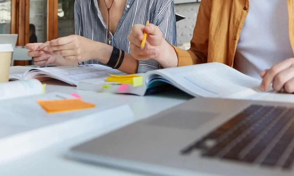Two people review documents and notes together at a desk, with sticky notes, open notebooks, and a laptop spread across the table.